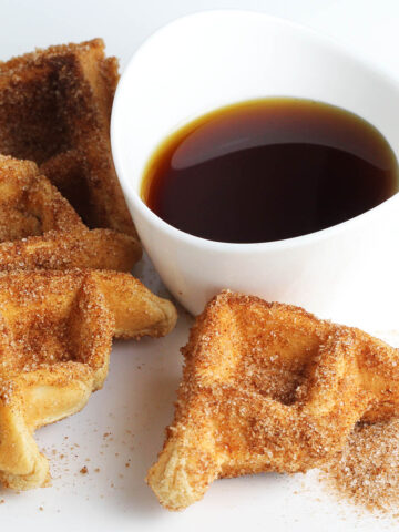 on the left a small waffle covered in cinnamon "sugar" (erythritol) with a couple smaller pieces around it, a small pile of cinnamon erythritol, and a small dipping bowl of maple syrup