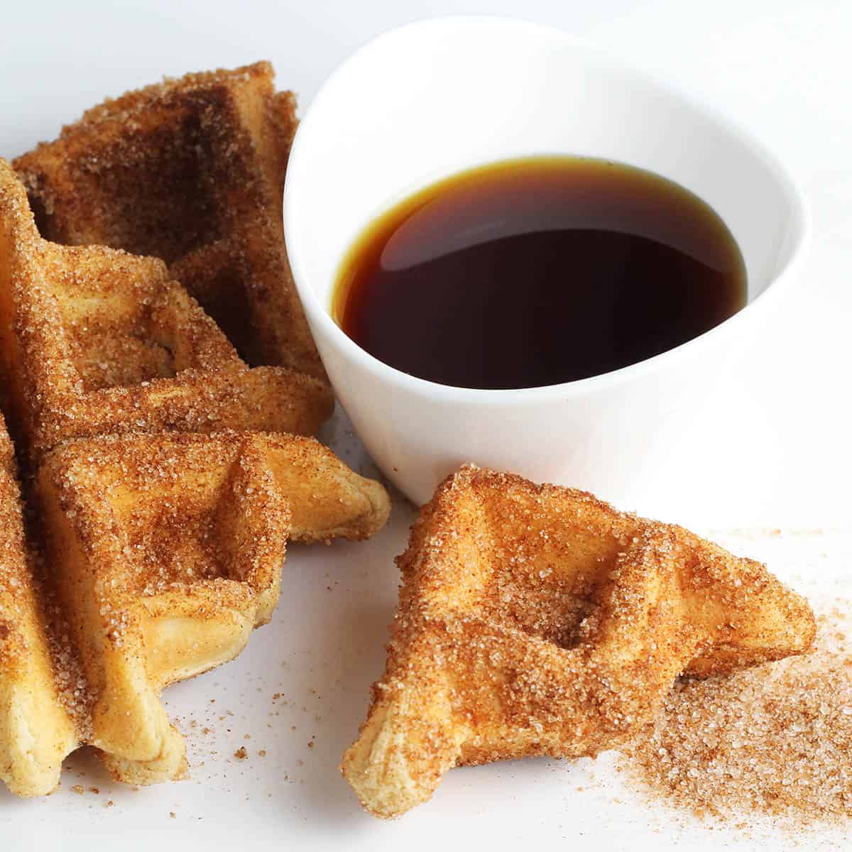 on the left a small waffle covered in cinnamon "sugar" (erythritol) with a couple smaller pieces around it, a small pile of cinnamon erythritol, and a small dipping bowl of maple syrup