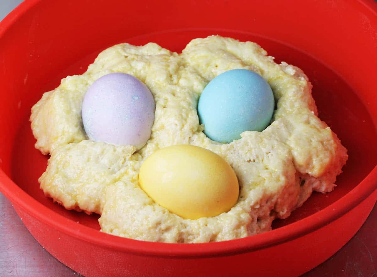 unbaked protein bread dough with colored easter eggs (purple, blue, and yellow) sitting into it all in a red round silicone cake pan