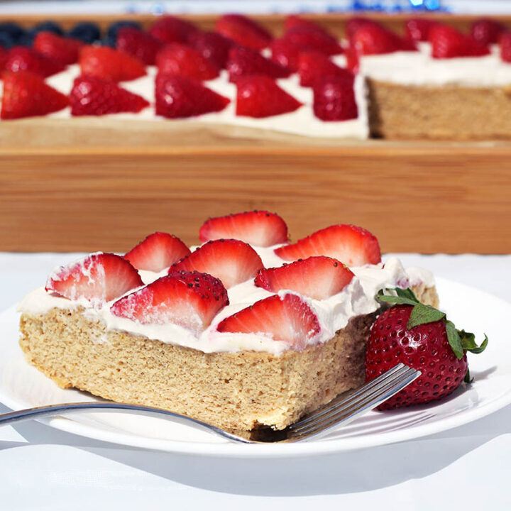square piece of vanilla protein cake topped with whipped cream and strawberries on a white plate with a fork and a whole strawberry