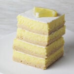 stack of protein lemon bars dusted with confectioners erythritol and a quarter of a lemon slice on top all on a white plate with a light background