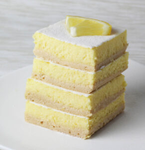 stack of protein lemon bars dusted with confectioners erythritol and a quarter of a lemon slice on top all on a white plate with a light background