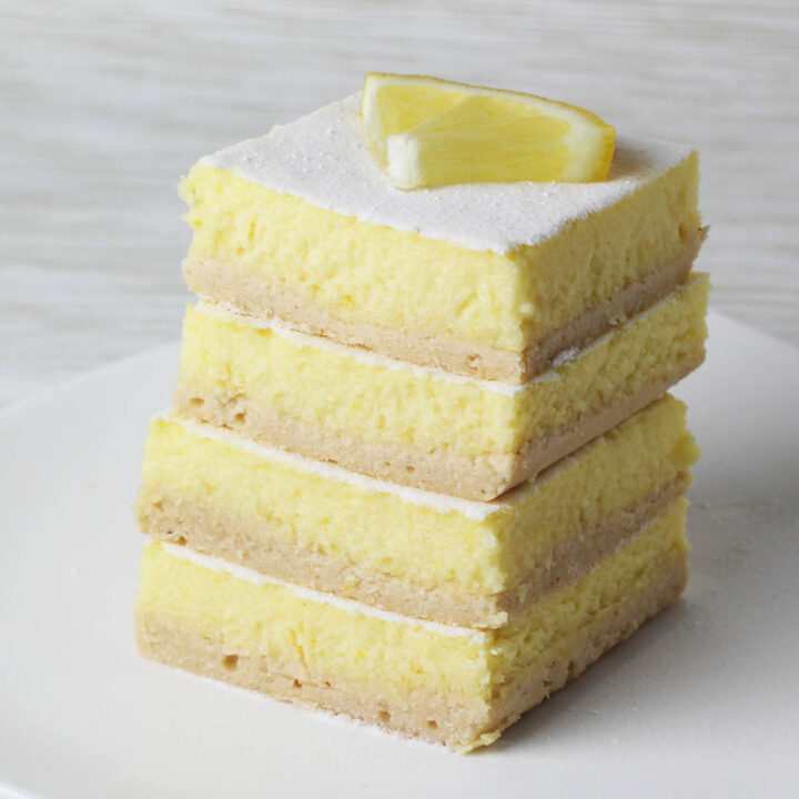 stack of protein lemon bars dusted with confectioners erythritol and a quarter of a lemon slice on top all on a white plate with a light background