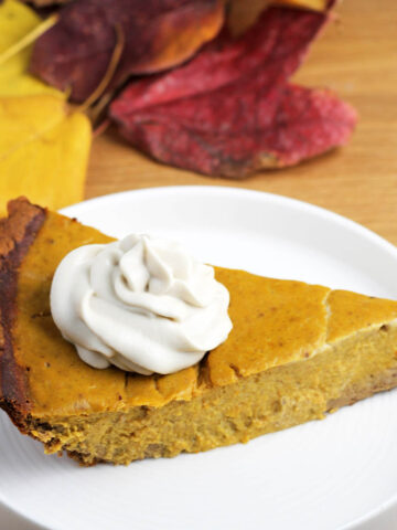 slice of protein pumpkin pie with a dollop of cashew whipped cream on top on a white plate on a wood table with fall colored leaves in the background
