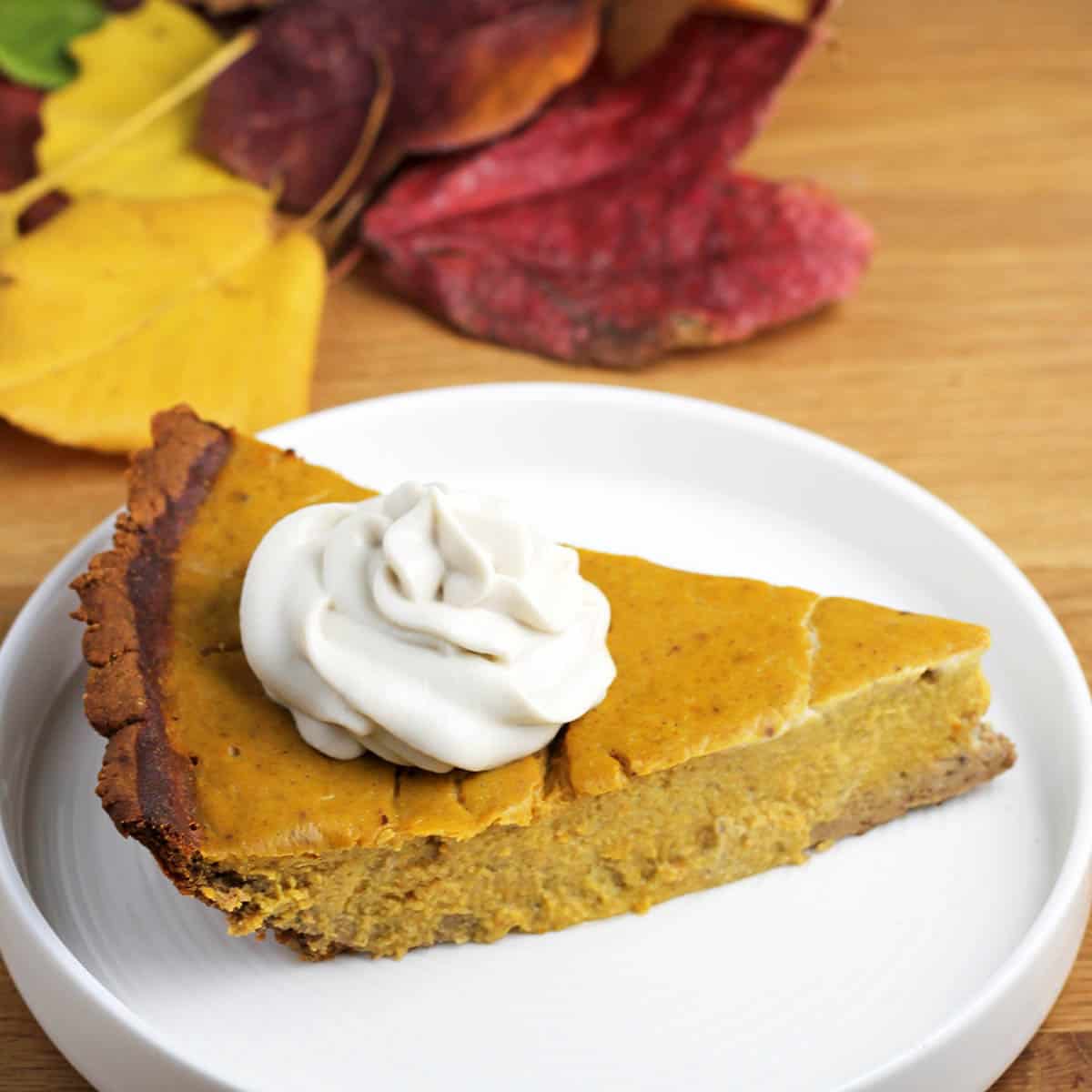 slice of protein pumpkin pie with a dollop of cashew whipped cream on top on a white plate on a wood table with fall colored leaves in the background