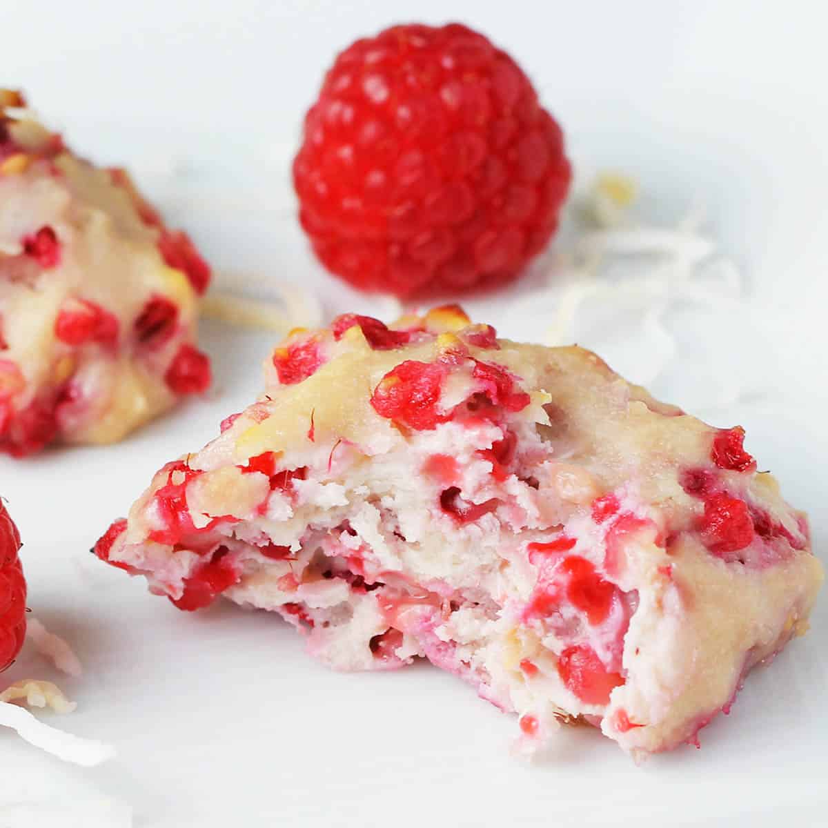 bitten coconut raspberry cookie - white cookie with bright raspberry pieces - with a fresh raspberry behind it all on a white plate