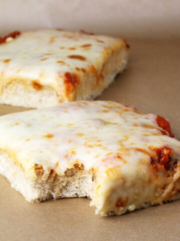 two slices of sicilian (square, thick crust) pizza on a sheet of unbleached parchment paper, a bite has been taken from the slice in the front