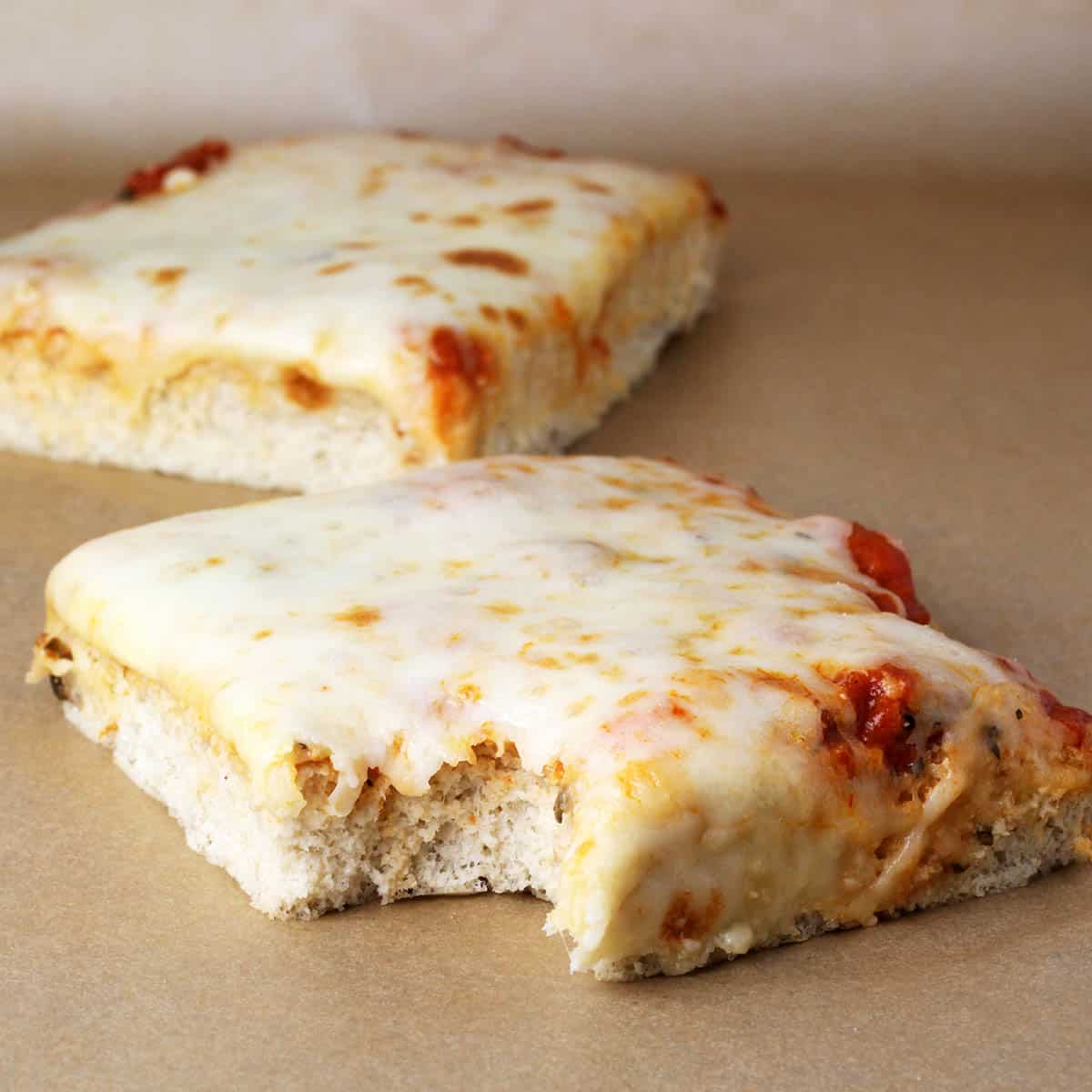 two slices of sicilian (square, thick crust) pizza on a sheet of unbleached parchment paper, a bite has been taken from the slice in the front