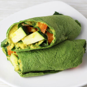 two halves of vegetable filled spinach protein wrap, one sitting diagonally on top of the other on a white plate