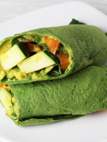 two halves of vegetable filled spinach protein wrap, one sitting diagonally on top of the other on a white plate