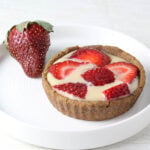a strawberries and cream tartlet with a whole strawberry on a round white plate on a light background