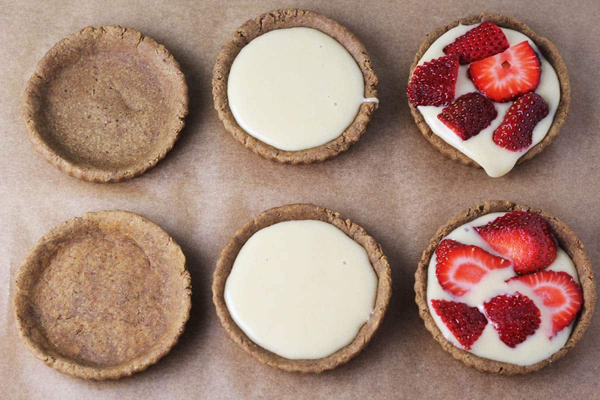 six mini tart shells, two are empty, two have vanilla whey cream filling, and two have vanilla whey cream with fresh strawberries on a sheet of unbleached parchment paper