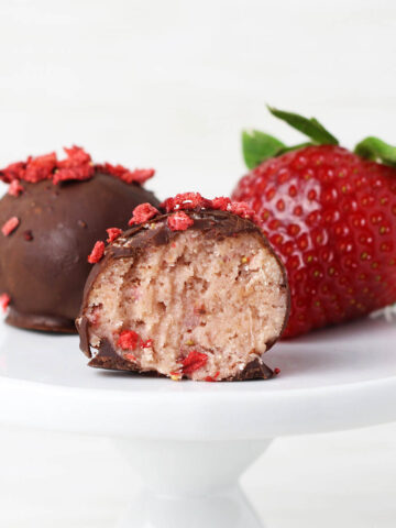 one full and one bitten chocolate covered strawberry protein ball next to a strawberry on a small white plate