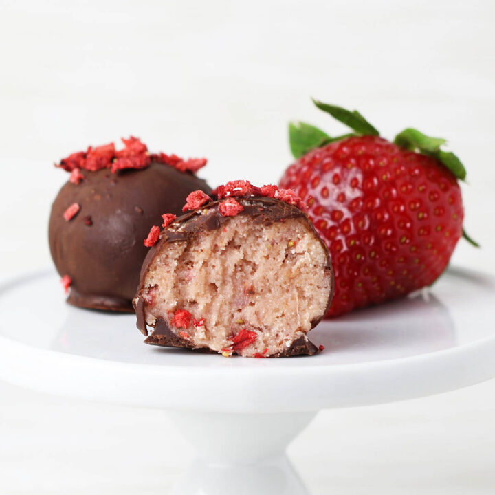 one full and one bitten chocolate covered strawberry protein ball next to a strawberry on a small white plate