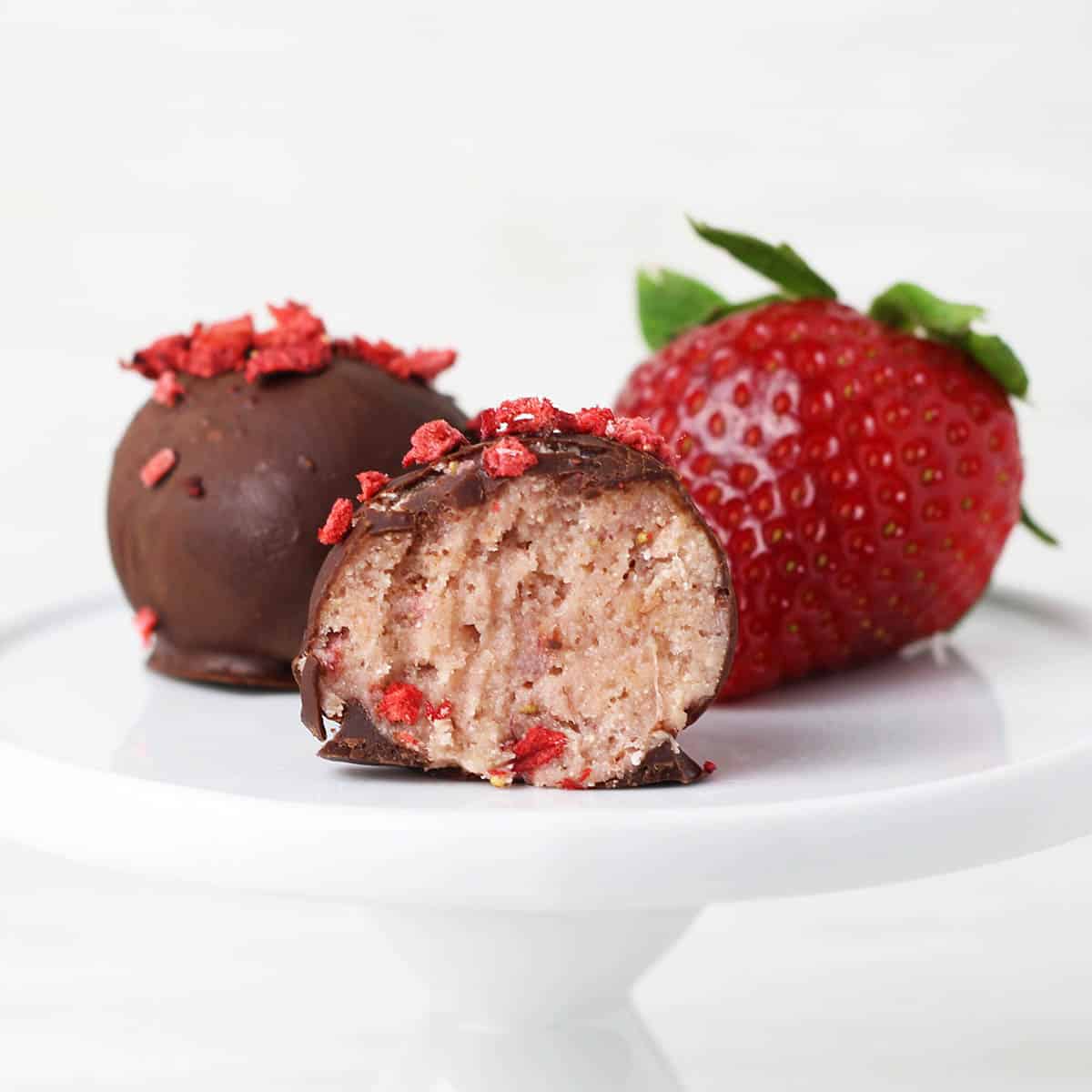 one full and one bitten chocolate covered strawberry protein ball next to a strawberry on a small white plate