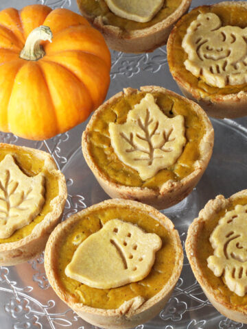seven mini pumpkin pies with fall shaped pie crust cookies on them next to a bright orange mini pumpkin on a flower etched glass cake stand