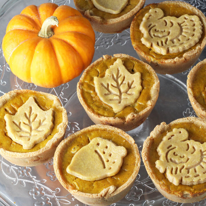 seven mini pumpkin pies with fall shaped pie crust cookies on them next to a bright orange mini pumpkin on a flower etched glass cake stand