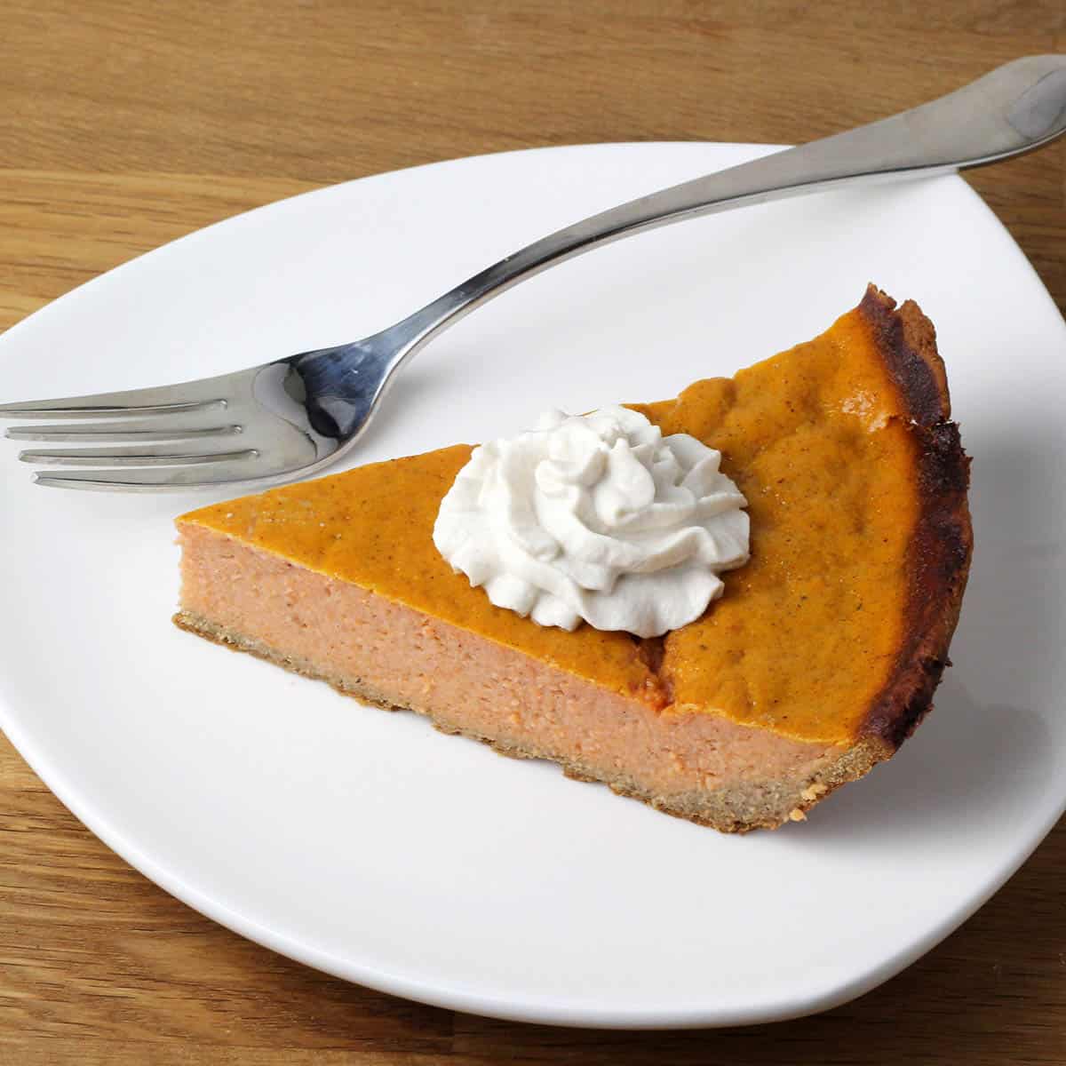 slice of sweet potato pie with a swirl of cashew whipped cream on a white plate with a silver fork on a brown wood table