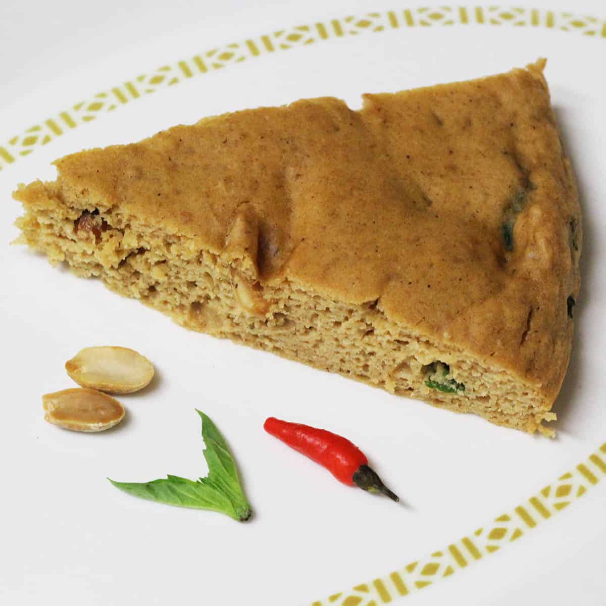 slice of thai peanut protein cake with a couple of peanut halves, a piece of thai basil, and a tiny red pepper on a white plate