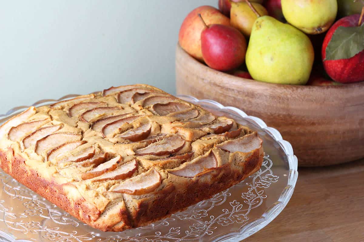 square vanill pear cake with sliced pears on the top on a round glass flower-etched cake stand, with a wooden bowl of pears in the background