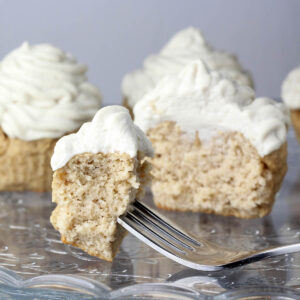 piece of frosted vanilla cupcake on a fork on an etched glass cake plate with cupcakes behind it.