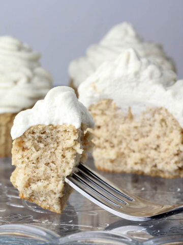 piece of frosted vanilla cupcake on a fork on an etched glass cake plate with cupcakes behind it.