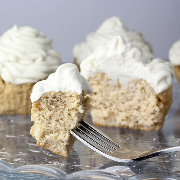 piece of frosted vanilla cupcake on a fork on an etched glass cake plate with cupcakes behind it.