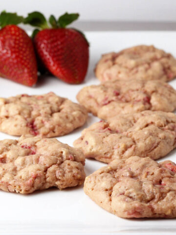 six cake-like strawberry protein cookies with visible strawberry pieces in them and two strawberries on a white plate