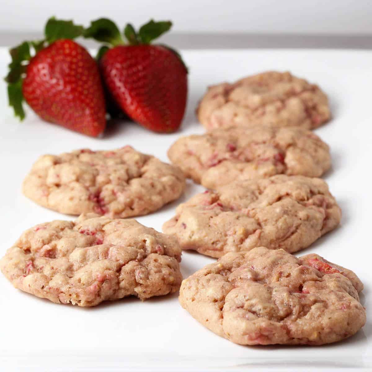 six cake-like strawberry protein cookies with visible strawberry pieces in them and two strawberries on a white plate