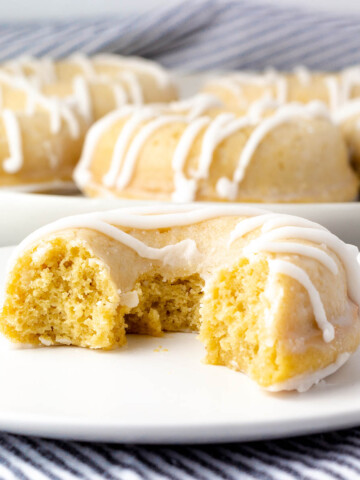 half of a glazed yellow cake donut with white drizzled icing on a white plate with a blue and white striped linen background and a plate with more donuts in the back