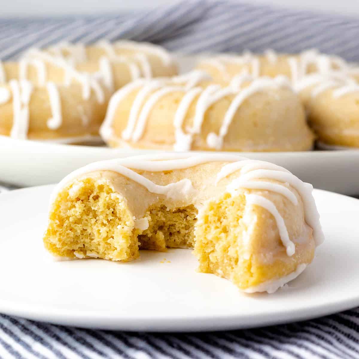 half of a glazed yellow cake donut with white drizzled icing on a white plate with a blue and white striped linen background and a plate with more donuts in the back