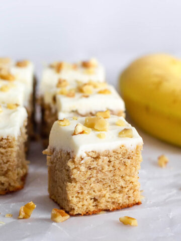 square pieces of frosted banana walnut cake with a ripe banana on the right and pieces of walnuts surround the cake pieces
