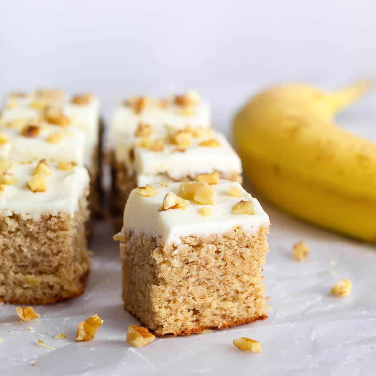 square pieces of frosted banana walnut cake with a ripe banana on the right and pieces of walnuts surround the cake pieces