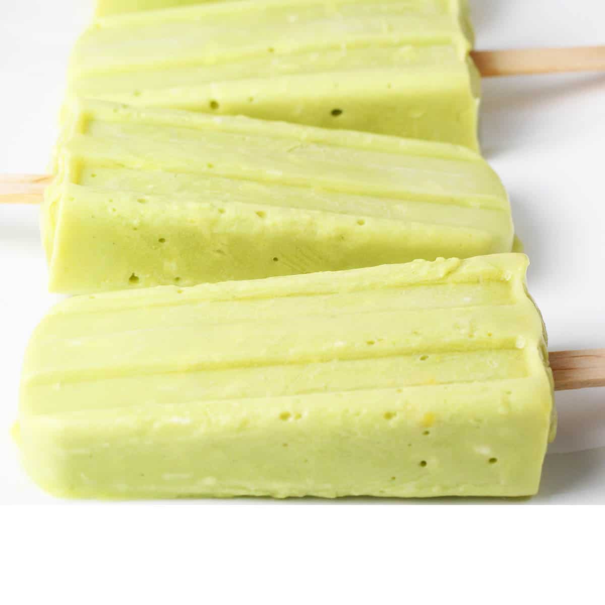 three coconut avocado popsicles sitting horizontally on a plate with the sticks alternating left and right