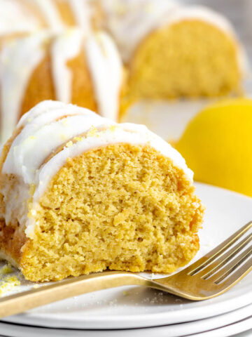 a slice of lemon-iced protein bundt cake on a plate with a gold form in front and the rest of the cake in the back and a lemon on the right