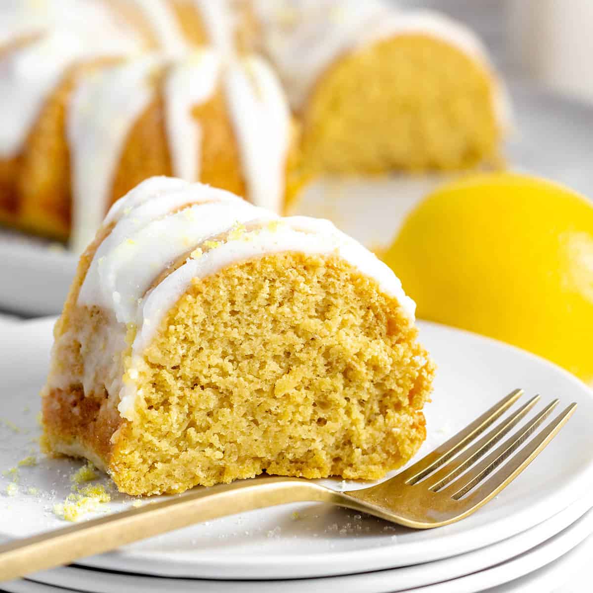 a slice of lemon-iced protein bundt cake on a plate with a gold form in front and the rest of the cake in the back and a lemon on the right