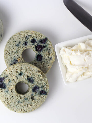 top view of a sliced blueberry bagel next to a small dish of cream cheese on a white plate with a butter knife