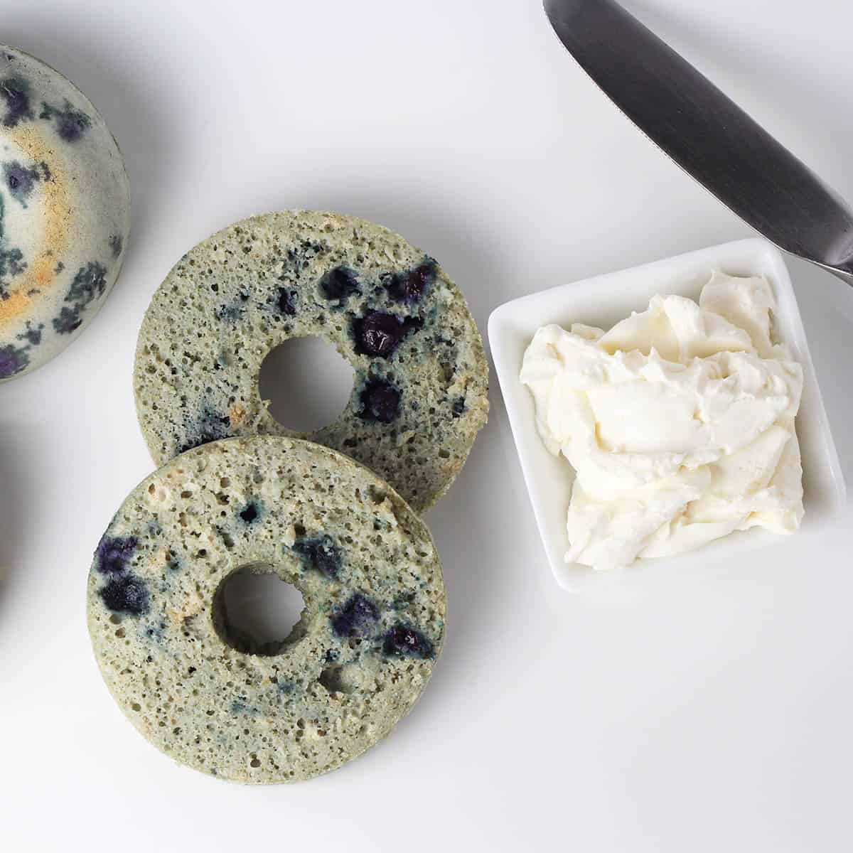 top view of a sliced blueberry bagel next to a small dish of cream cheese on a white plate with a butter knife