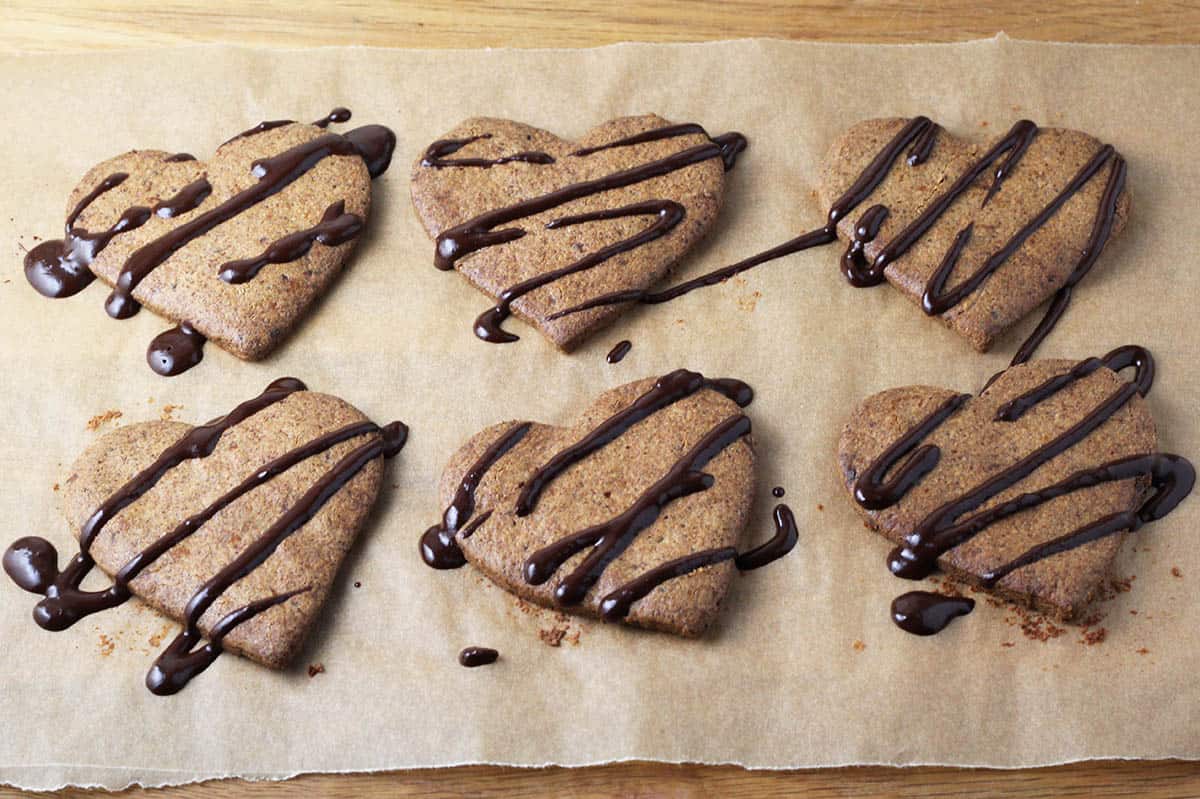 six baked heart shaped almond cookies with fresh chocolate drizzle on a piece of unbleached parchment paper