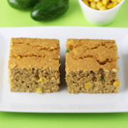 two square pieces of protein corn bread with visible pieces of corn and jalapeno on a white plate with two jalapenos and a small dish of corn behind it