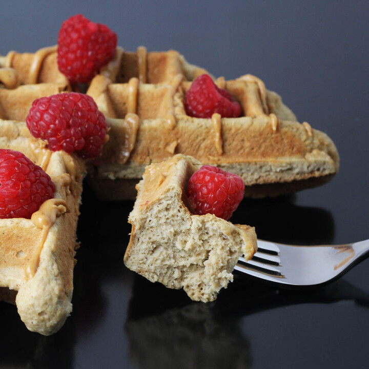 three quarters of a peanut butter belgian waffle topped with raspberries, and a fork in front with a bite of waffle and a raspberry