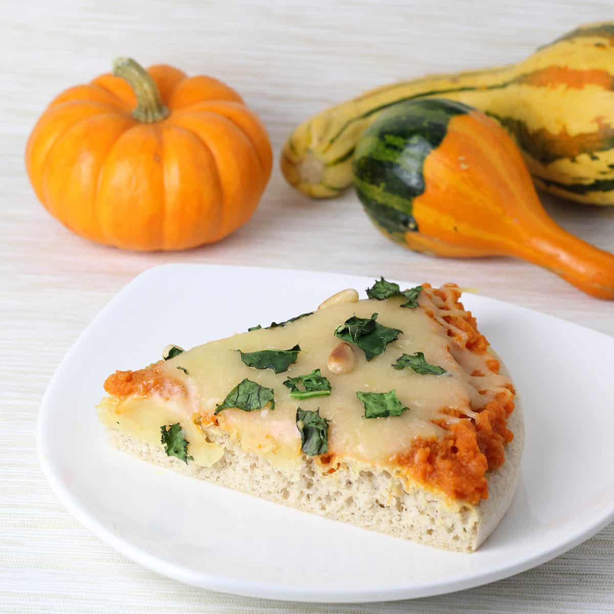 slice of pumpkin kale pizza on a white plate an next to a mini pumpkin and mini squash