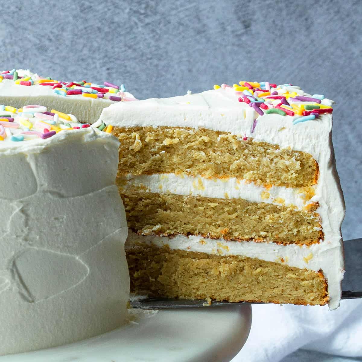 slice of frosted 3 layer cake being pulled out with a cake server, with a grey background