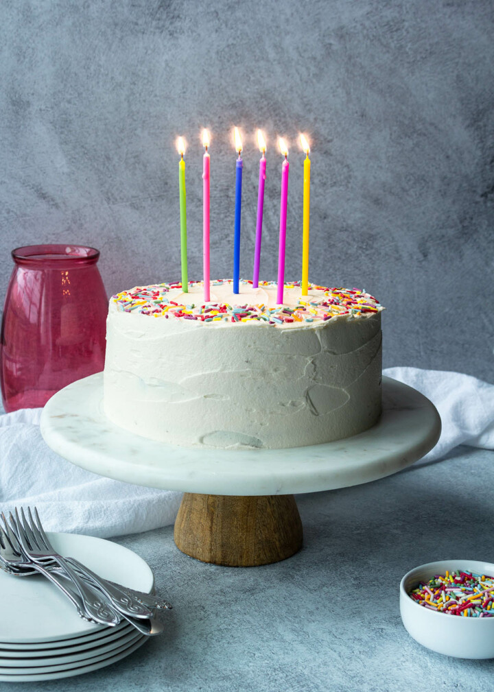 protein birthday layer cake with vanilla frosting, naturally colored sprinkles, brightly colored tall candles that are lit, on a marble cake stand with a wooden base. also in view are a stack of plates with forks on top, a small dish of sprinkles, and a pink glass vase in the background