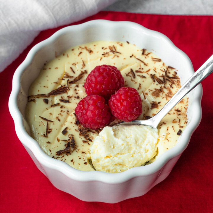 ramekin of protein cheesecake with chocolate shavings and three raspberries on top and a spoonful showing the creamy texture, on a red placemat