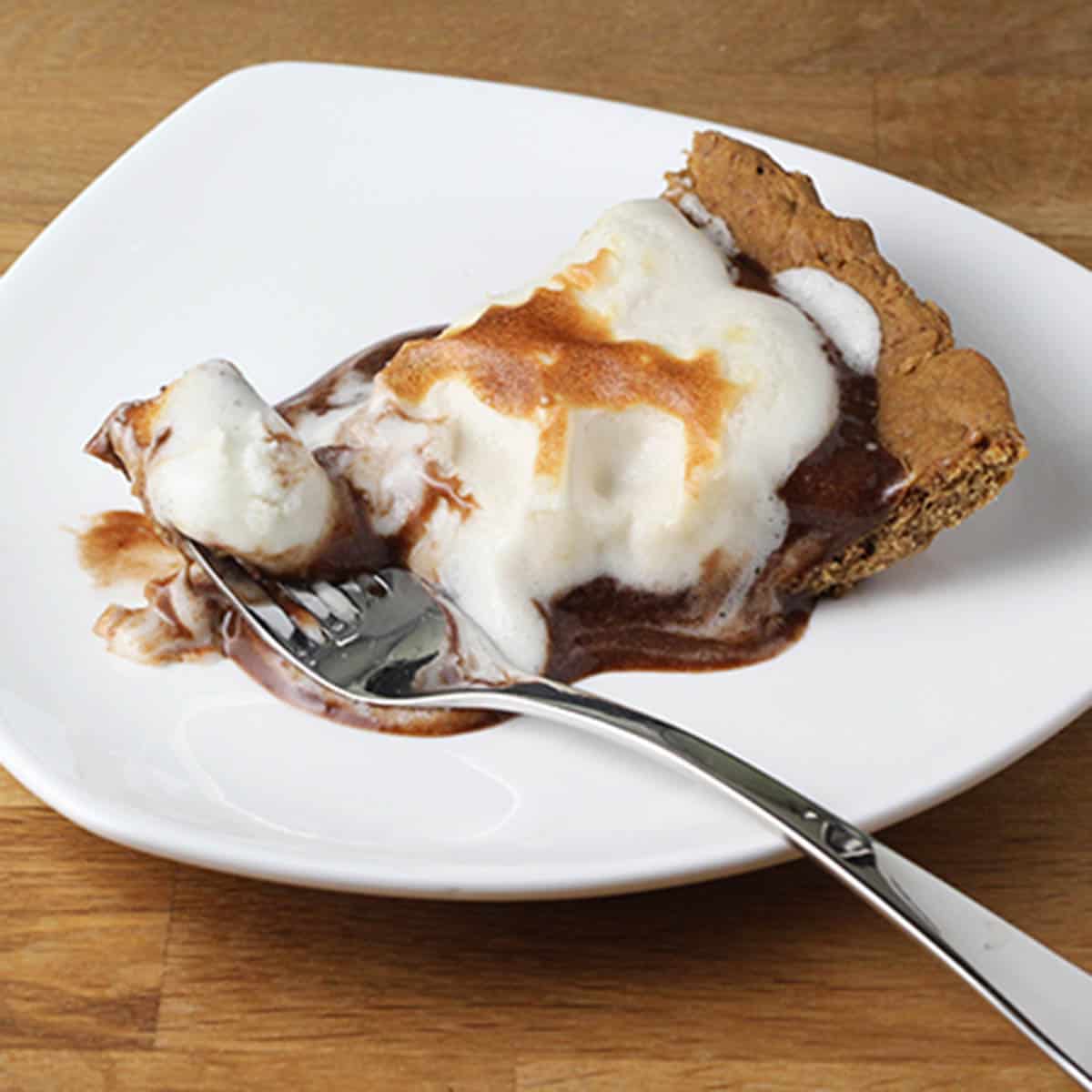 a piece of s'mores protein pie - cholate filling topped with toasted protein marshmallow fluff - on a white plate with a silver fork on a wood table