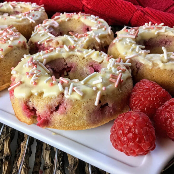 several raspberry donuts topped with pink sprinkles on a plate with 3 fresh raspberries