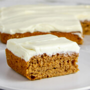 square piece of pumpkin snack cake with cream cheese frosting on a white plate in front of the rest of the cake