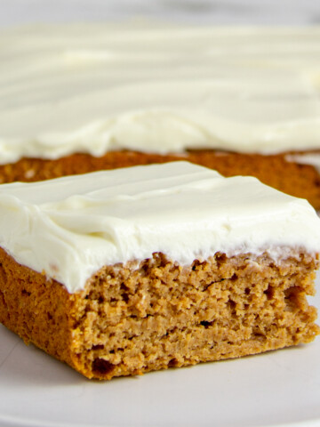square piece of pumpkin snack cake with cream cheese frosting on a white plate in front of the rest of the cake
