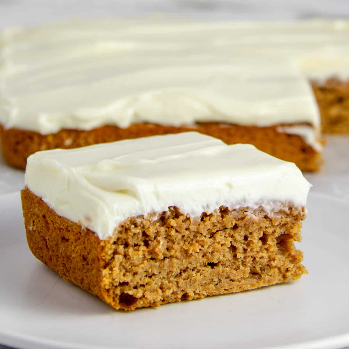 square piece of pumpkin snack cake with cream cheese frosting on a white plate in front of the rest of the cake
